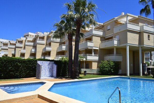 A swimming pool with a geometric tile pattern, surrounded by a landscaped area with palm trees and a building with multiple balconies. - Ap2236 in Denia With 2 Bedrooms and 2 Bathrooms (Denia)