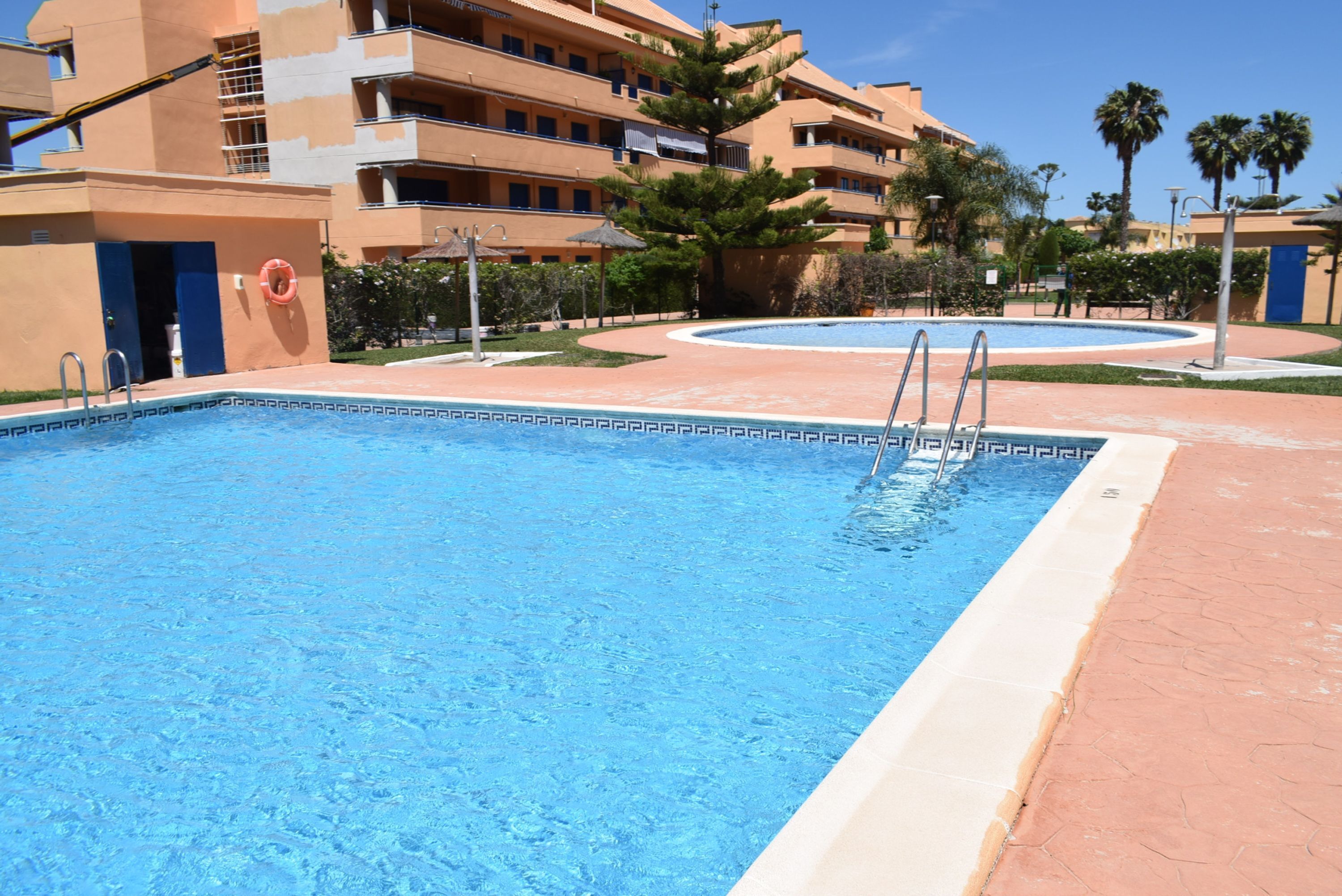 Una piscina con agua cristalina azul, rodeada por un paseo de baldosas y un edificio con varios balcones.