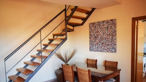 Un comedor con una mesa y sillas de madera, una escalera con barandilla de metal, y una pared decorada con una obra de arte estampada.
