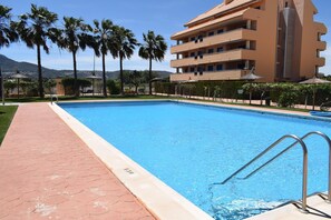 A swimming pool with a tiled deck, surrounded by palm trees and a multi-story building with balconies. - At2214 in Denia With 2 Bedrooms and 2 Bathrooms (Denia)