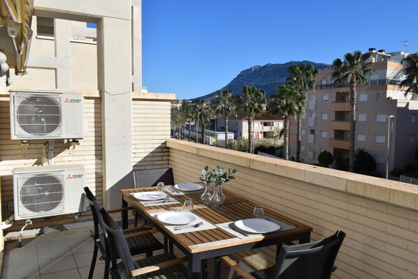 Un balcon avec une table en bois mise pour les repas, des unités de climatisation et une vue sur les palmiers et les immeubles.