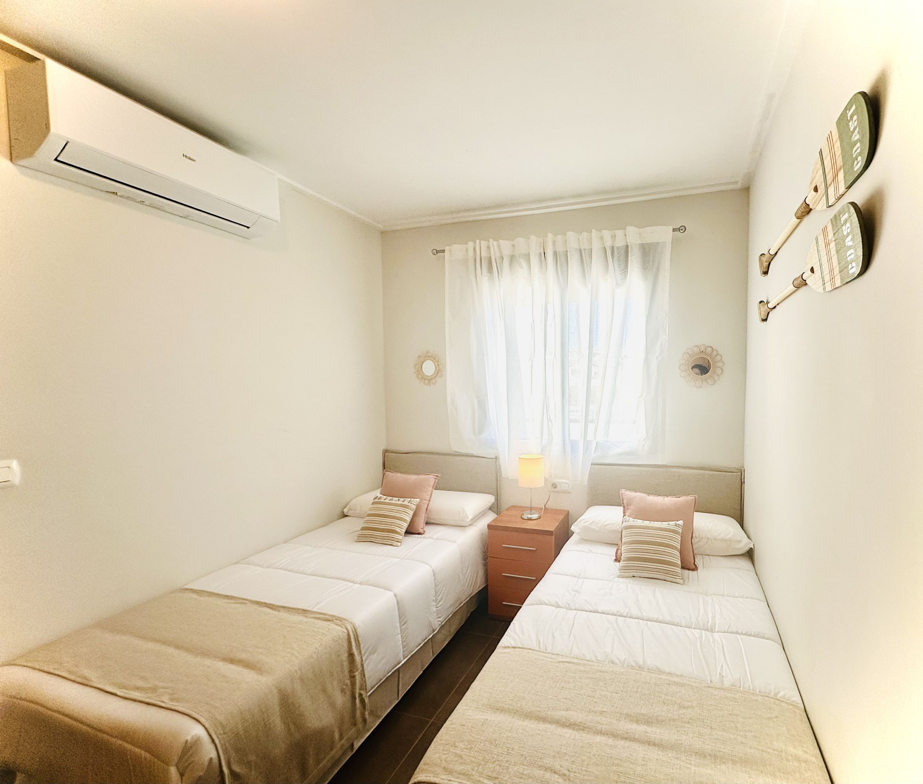 Une chambre avec deux lits, une table de chevet avec une lampe et une fenêtre avec des rideaux légers.