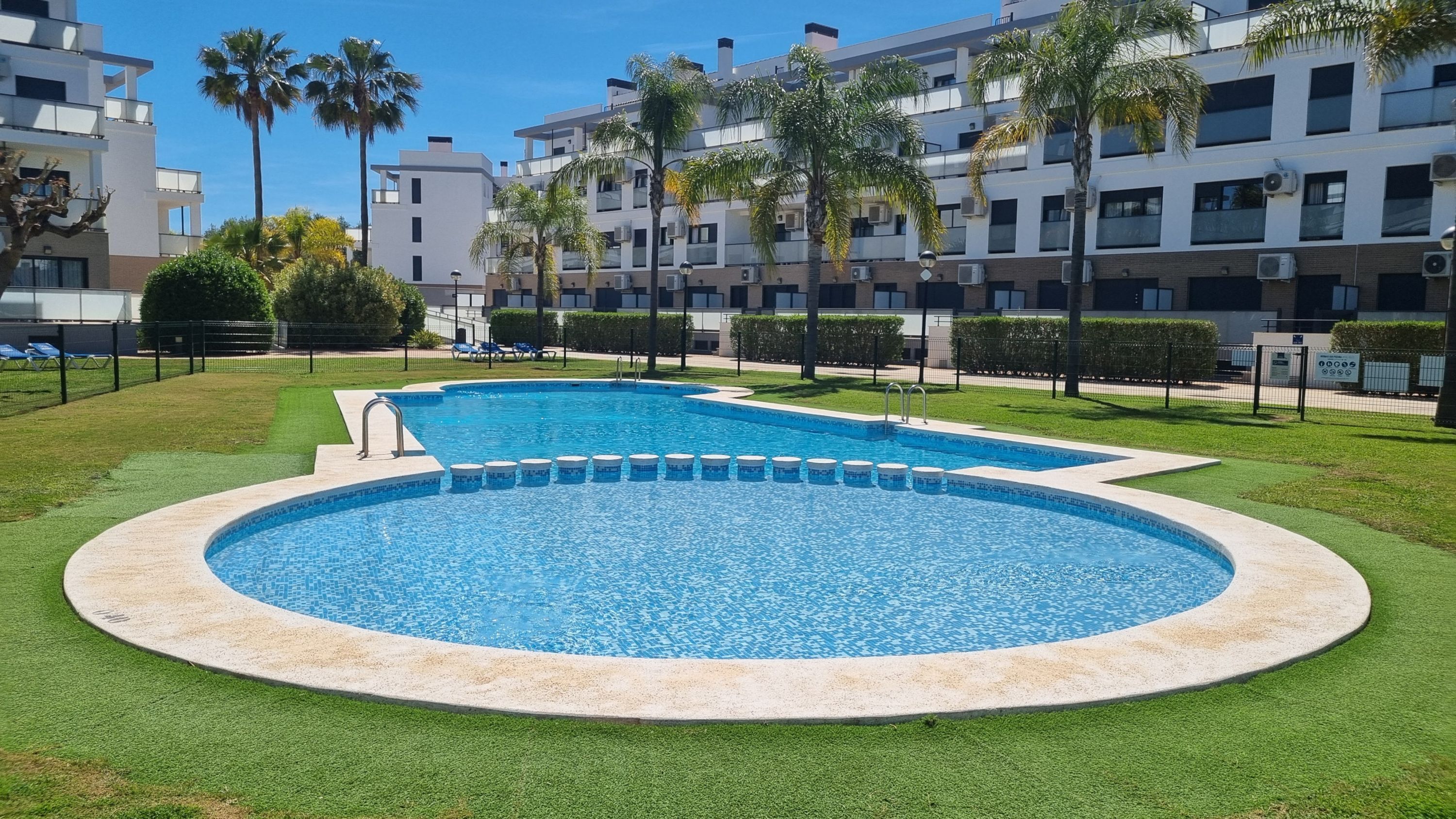En pool med cirkelformad design, omgiven av välskött gräsmatta och palmer, med moderna byggnader i bakgrunden.
