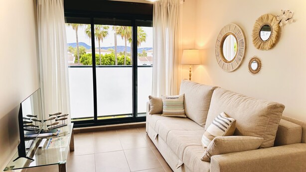 A living room with a beige sofa, a glass coffee table, and a view of palm trees and mountains through large windows. - Ap313 in Oliva With 2 Bedrooms and 1 Bathrooms (Oliva)