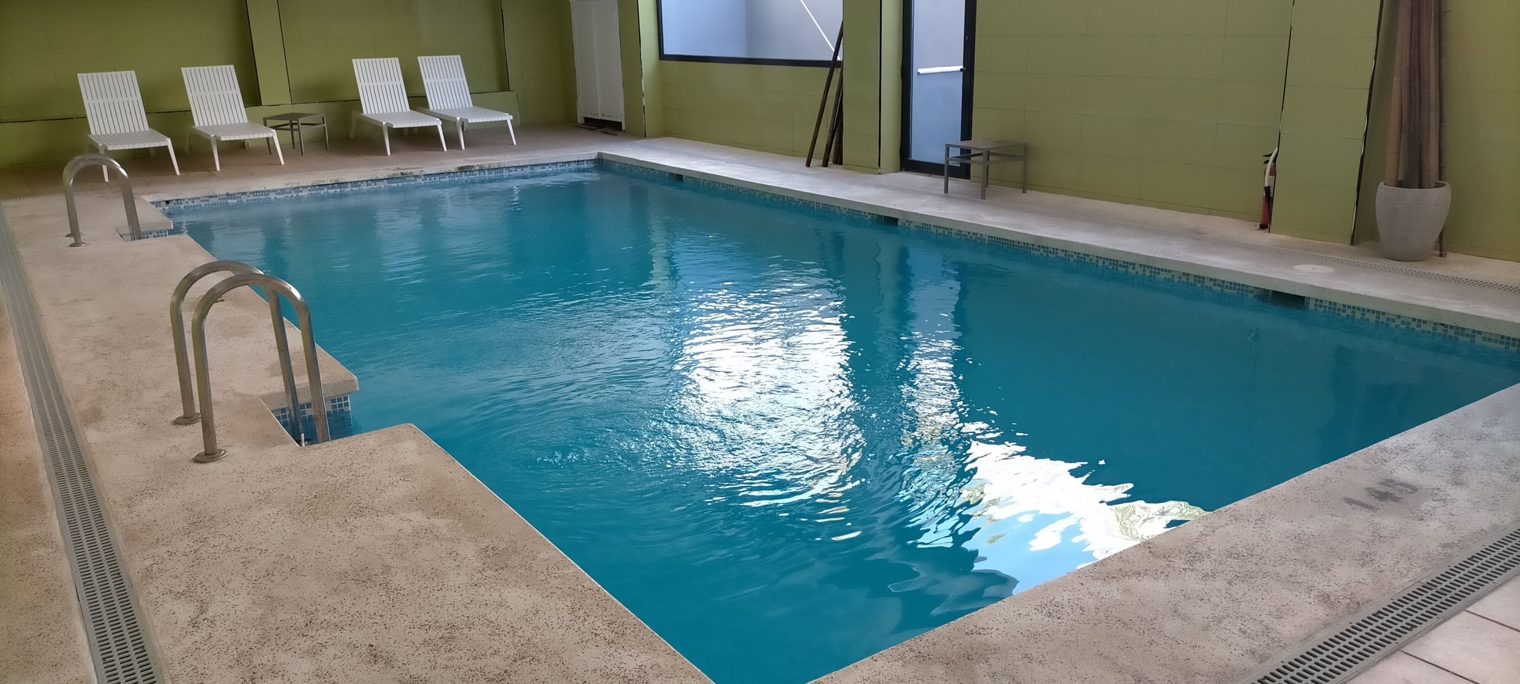 Una piscina interior con agua azul cristalina, rodeada de sillones blancos y un suelo de baldosas.