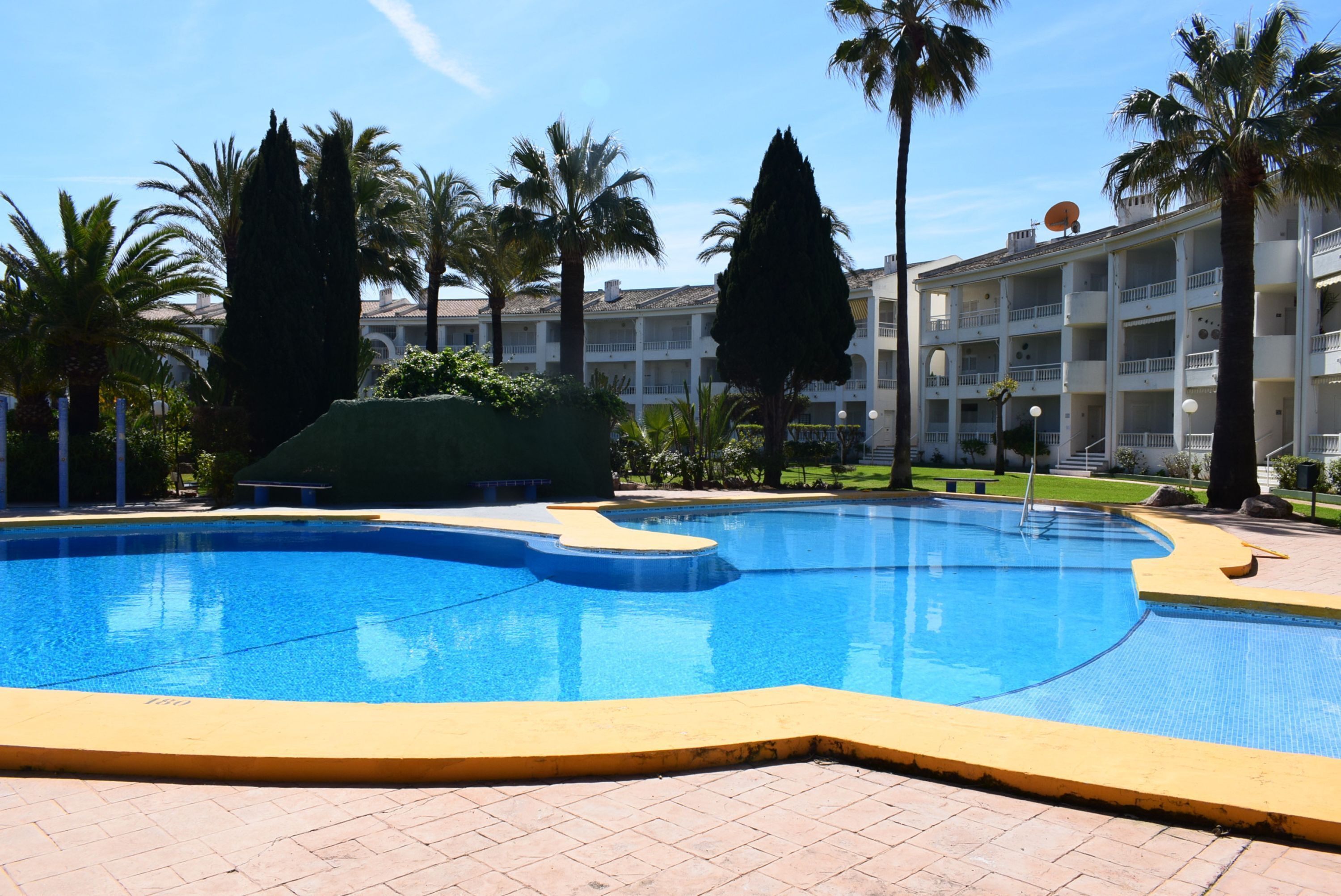 A swimming pool with a yellow border, surrounded by palm trees and a building with balconies.