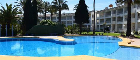 A swimming pool with a yellow border, surrounded by palm trees and a building with balconies.
