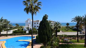 A pool area with a clear view of the sea, surrounded by palm trees and a building with a tiled roof.