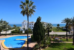 Zona de piscina con vista despejada al mar, rodeada de palmeras y un edificio con techo de tejas.