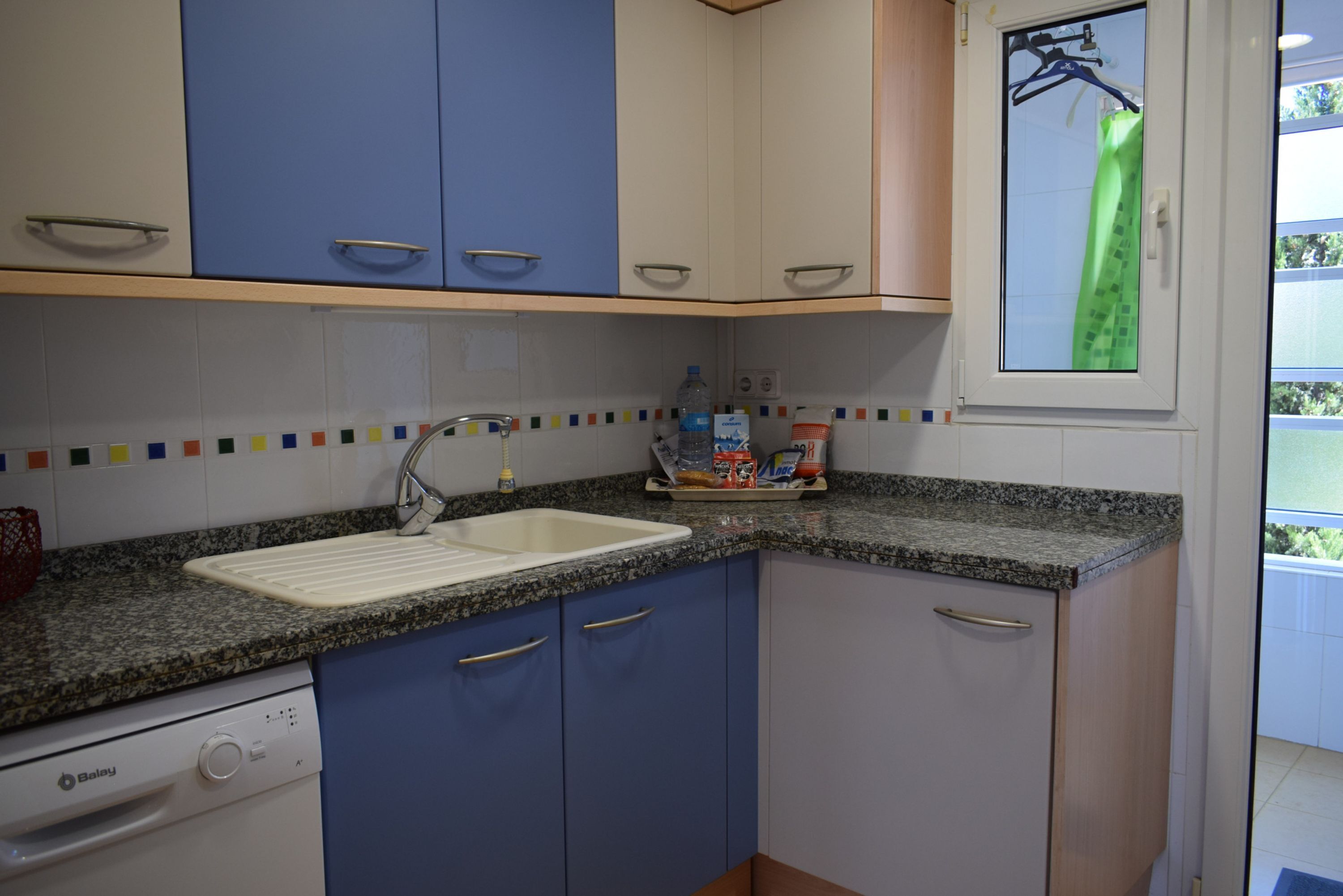 Eine Küche mit blauen und weißen Schränken, einer Granitarbeitsplatte, einem Geschirrspüler und einem Fenster mit Blick auf Grünflächen.