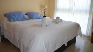 A neatly made bed with white bedding and blue pillows, a bedside lamp, and a window with sheer curtains. - Ap2215 in Denia With 2 Bedrooms and 2 Bathrooms (Denia)