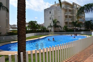 Ein Schwimmbecken, umgeben von einem weißen Zaun, mit Palmen und einem mehrstöckigen Gebäude im Hintergrund.