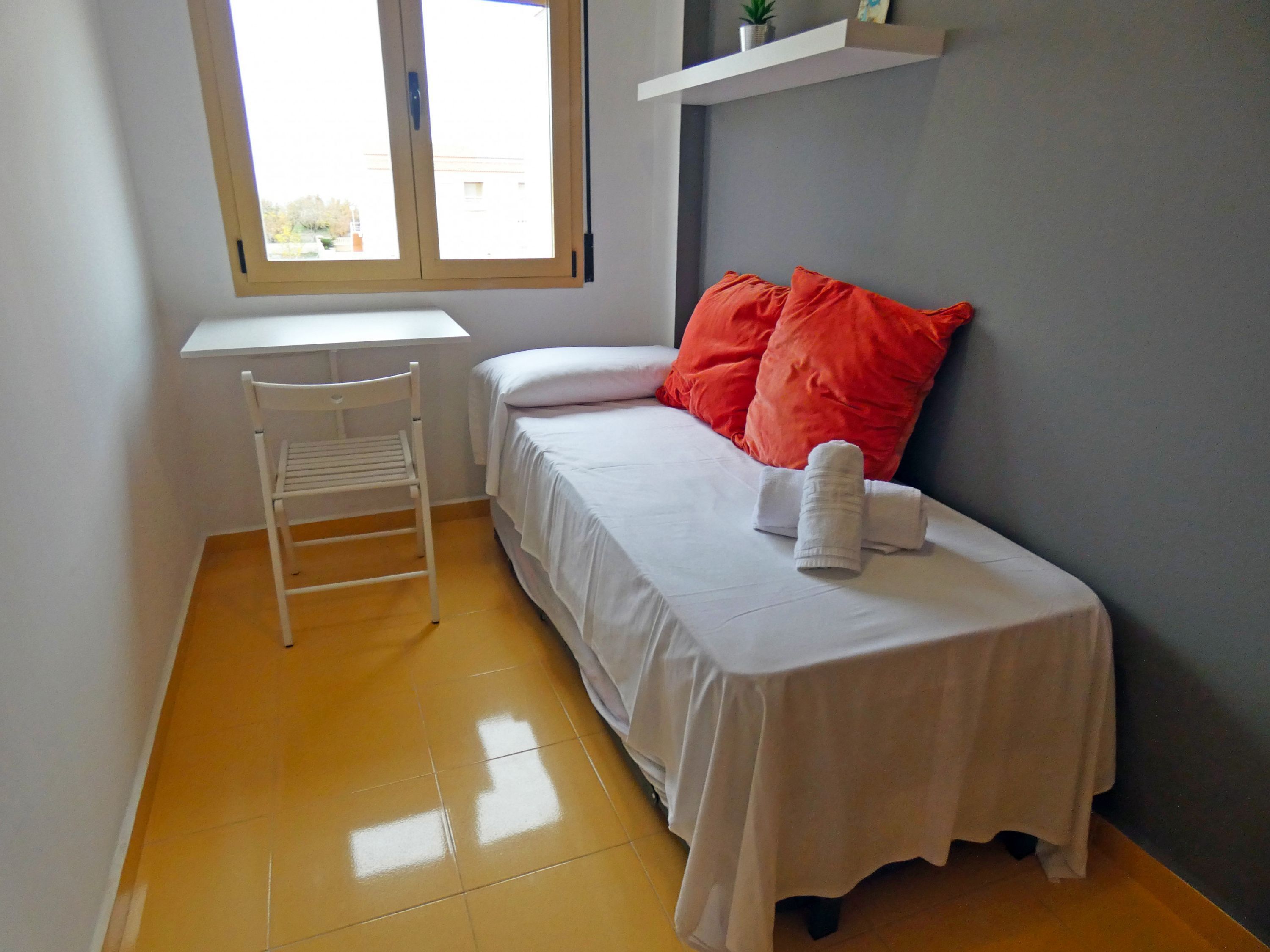 A small, tidy room with a bed, a white chair, a small table, and a window.