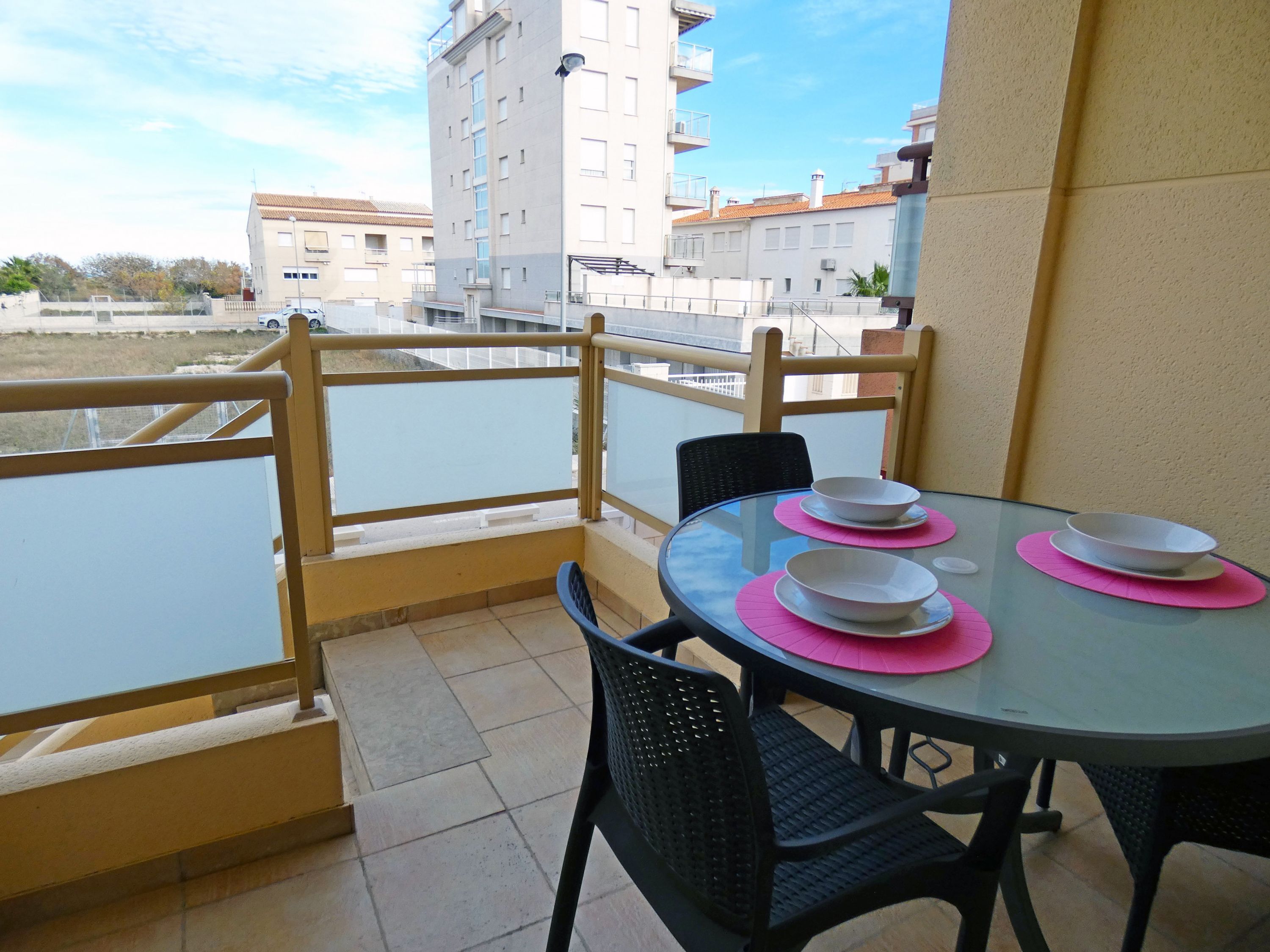 Een balkon met een glazen tafel, voorzien van witte schalen en roze placemats, omringd door zwarte stoelen.