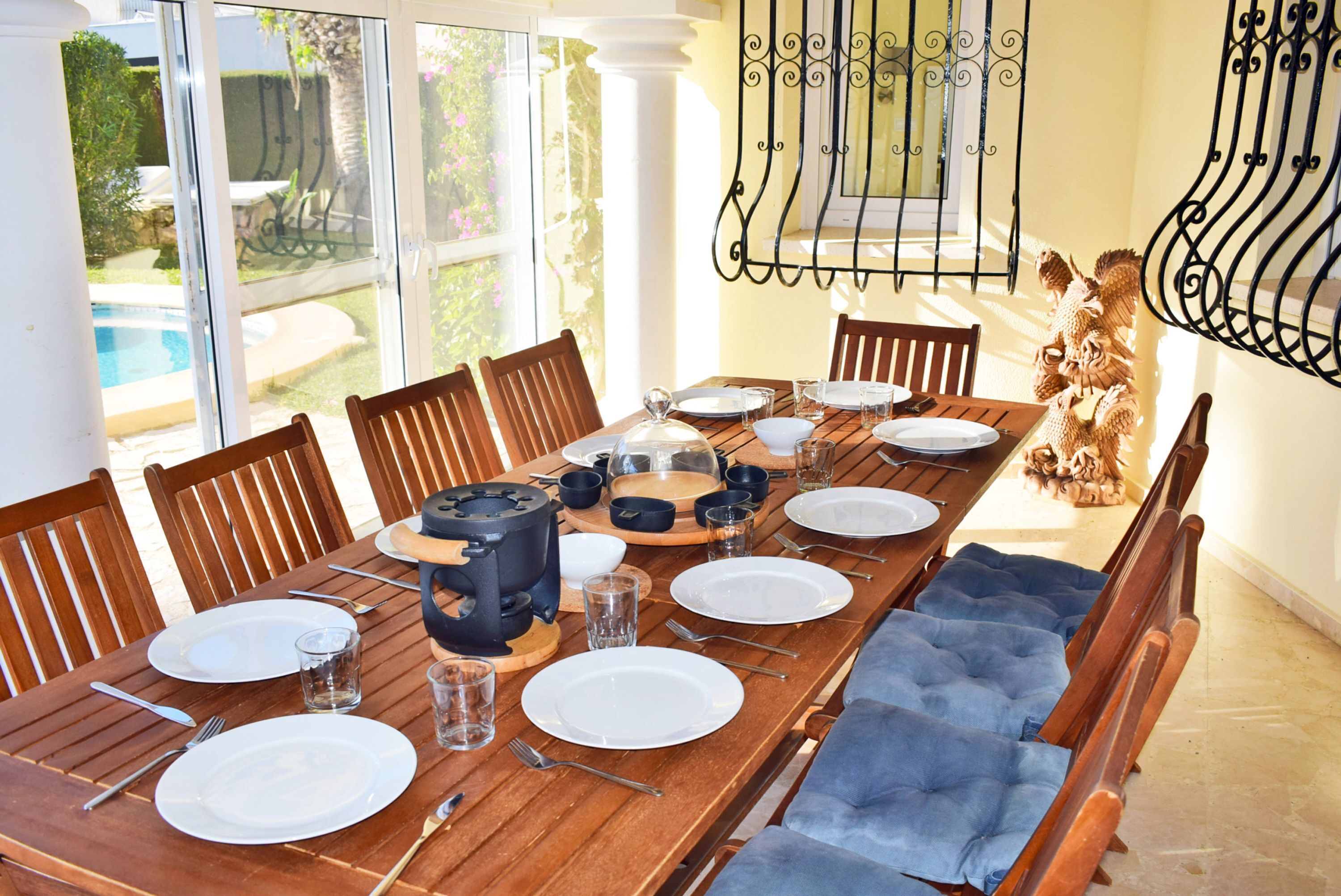 A dining area with a long wooden table set for a meal, surrounded by chairs, with a view of a swimming pool through large windows.