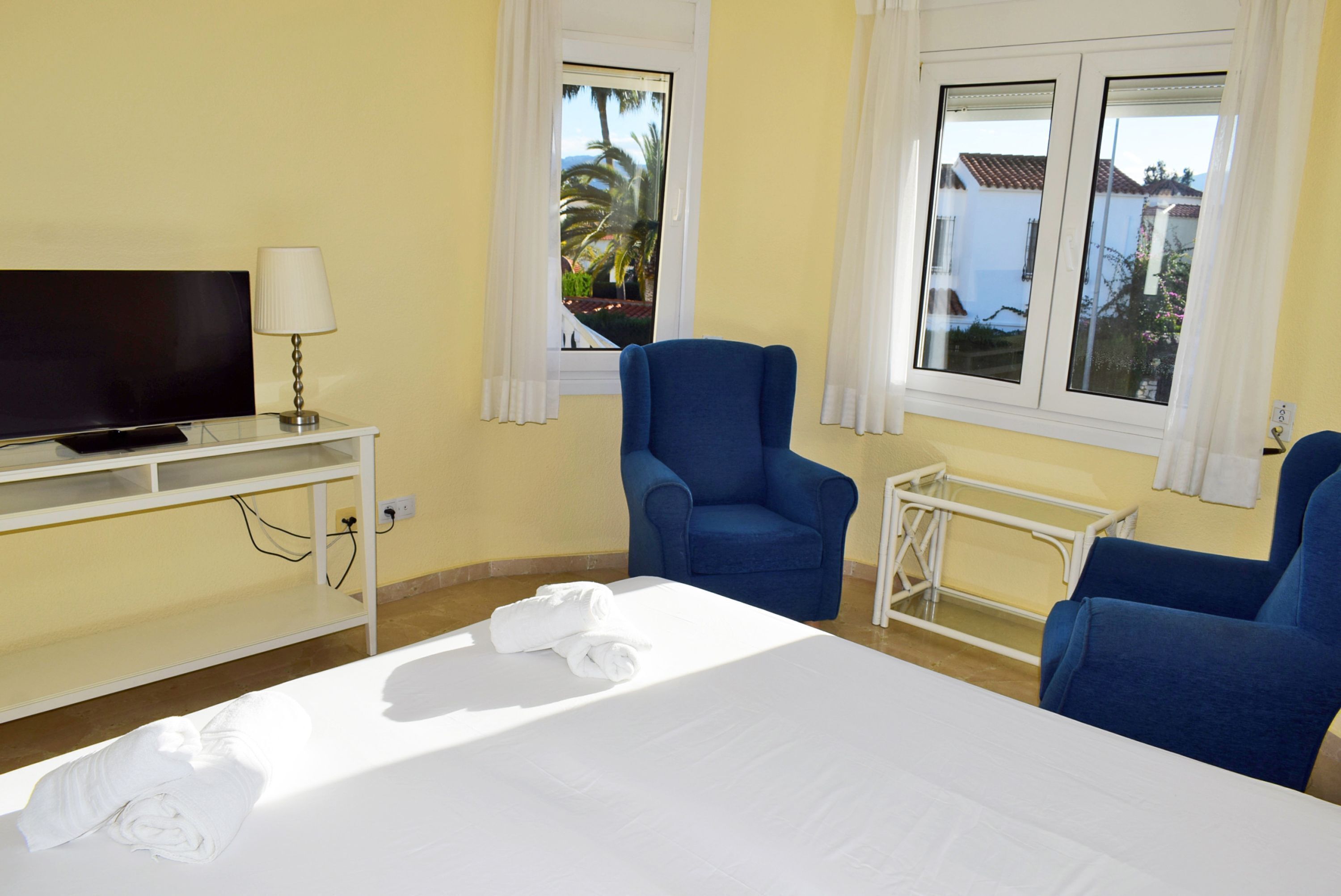 A hotel room with a bed, a TV, a desk with a lamp, and two blue armchairs.