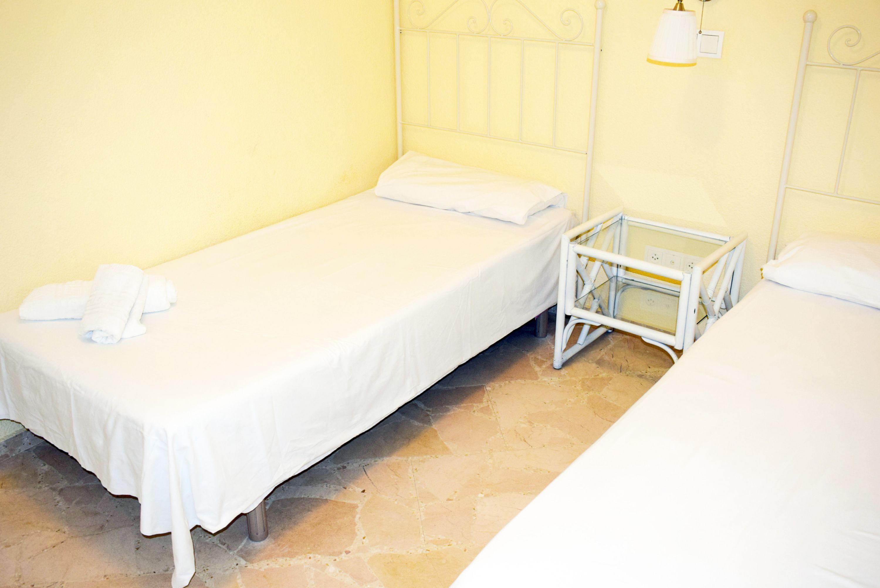 Une chambre avec un lit simple, un cadre de lit en métal, une table de chevet avec une lampe et un mur jaune uni.