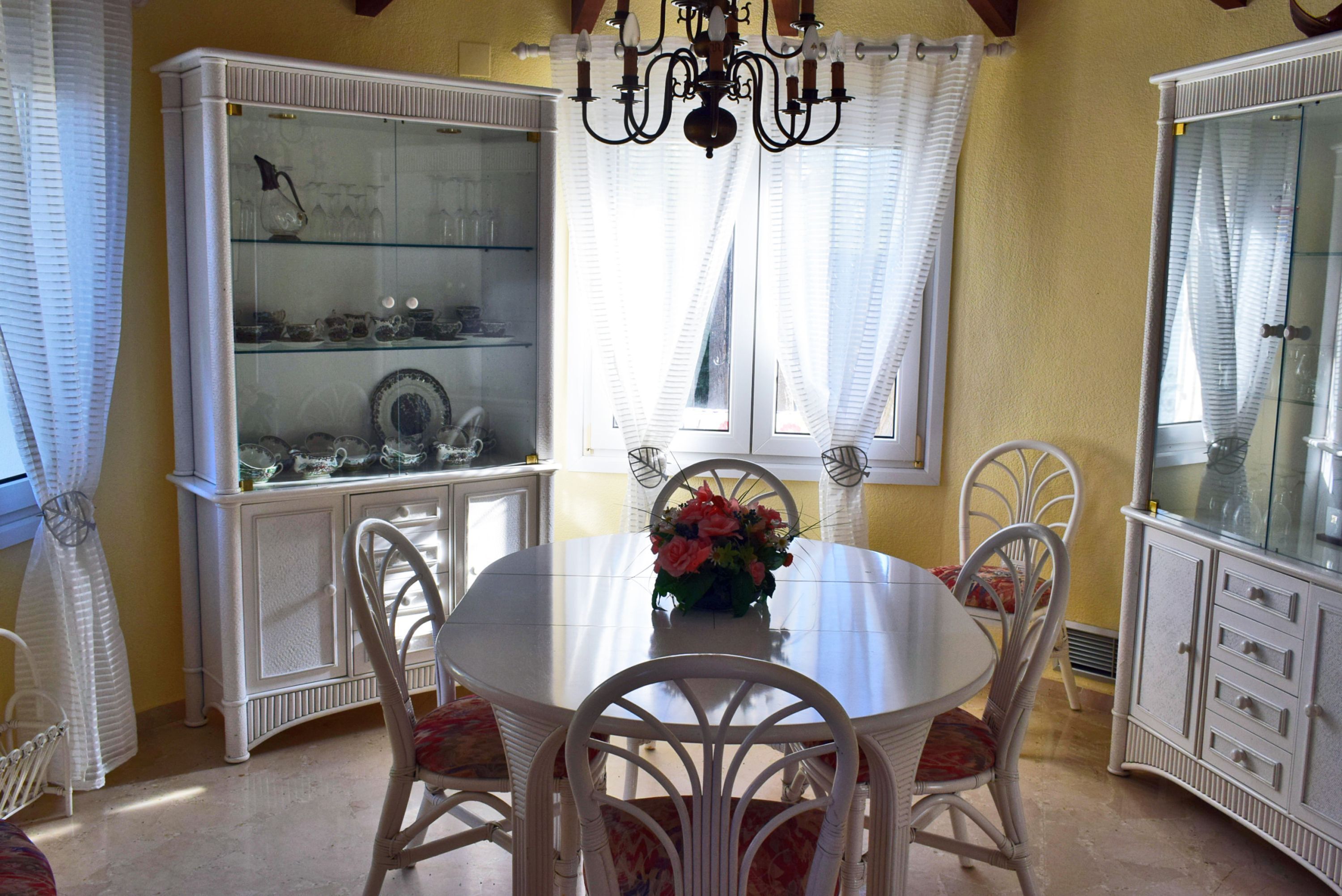 Un comedor con una mesa redonda, sillas blancas, una araña y armarios de vidrio con vajilla.