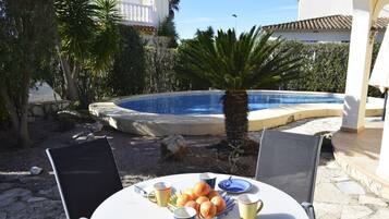 Une table placée au bord de la piscine, avec des fruits et des gobelets, entourée de chaises, avec une piscine et des palmiers en arrière-plan.