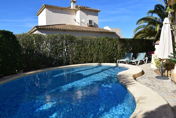 A swimming pool with a curved edge, a white building with a tiled roof in the background, and a palm tree to the right. - Ch1400 in Oliva With 2 Bedrooms and 2 Bathrooms (Oliva)