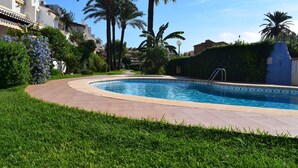 A swimming pool with a curved edge, surrounded by a well-maintained lawn and palm trees. - Dup2136 in Denia With 2 Bedrooms and 1 Bathrooms (Denia)