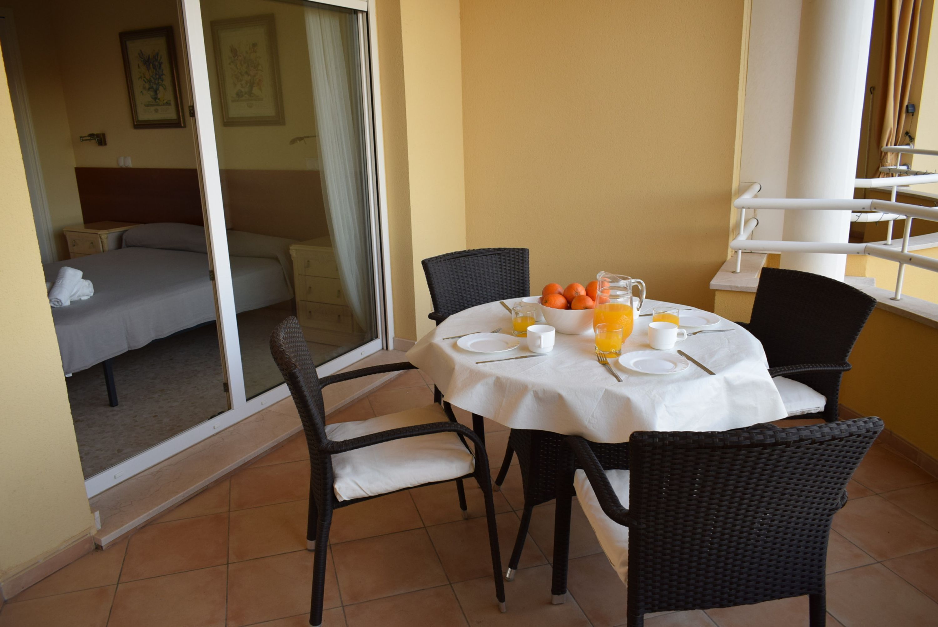 Un área de comedor con una mesa preparada para dos personas, una silla de mimbre y vistas a un dormitorio a través de una puerta de vidrio.
