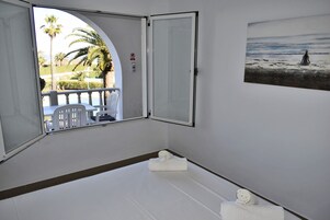 Una habitación de hotel con un ventanal grande, una cama blanca y un cuadro con una playa.
