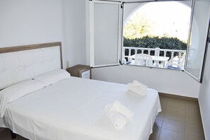 Un lit bien fait, avec du linge de lit blanc et deux serviettes enroulées, une petite table de chevet et une fenêtre donnant sur un balcon blanc et une végétation luxuriante.