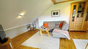 Una acogedora sala de estar estilo loft con piso de madera, un sofá gris con cojines naranja, una mesa de centro de madera y un mueble de madera con puertas de vidrio.
