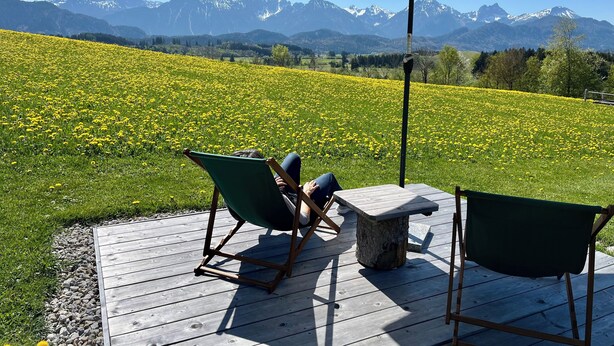 Una terraza de madera con dos sillas plegables verdes y una mesa de piedra, con vistas a un campo de flores amarillas y una cordillera.