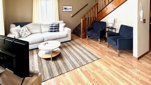 Un salón espacioso con un sofá blanco, dos sillones azules, una escalera de madera y un televisor sobre un mueble de madera.