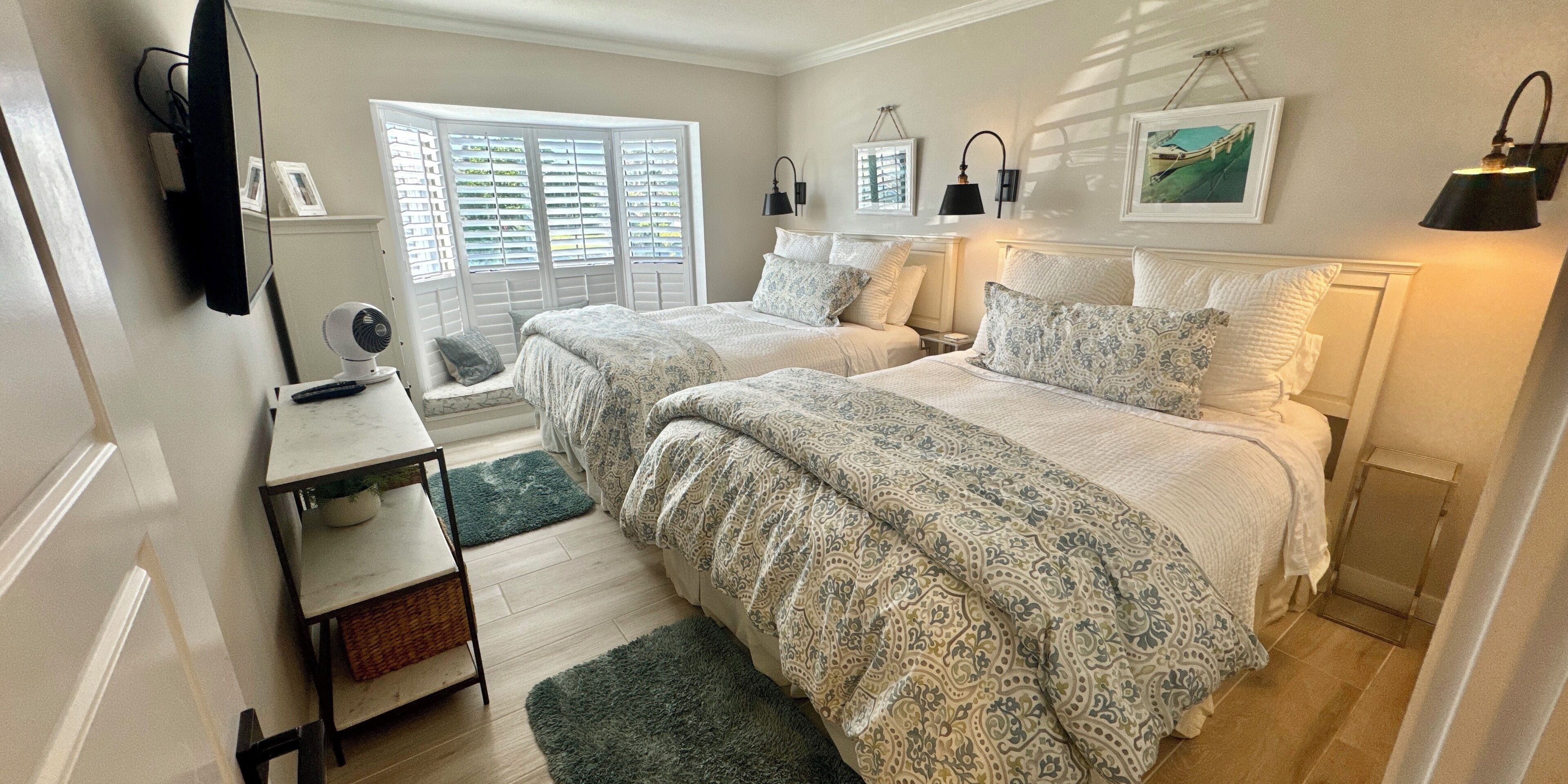 A bedroom with two beds, a desk, a chair, a standing fan, and a window with shutters.