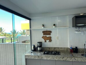 A kitchen with a granite countertop, a coffee maker, a gas stove, and a sign that reads "SEJAM BEM VINDOS CANTINHO DO CHURRASCO". - Flat Premium Vista Mar - Praia de Tabatinga (Conde)