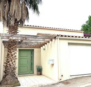 Un edificio de una sola planta con una puerta verde, una planta pequeña en maceta y una pérgola de madera.