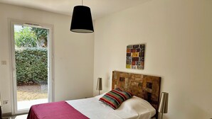 Un dormitorio con cama, colcha roja, cabecera de madera, un cuadro con una placa de matrícula en la pared, mesita de noche con lámpara y una silla de mimbre.