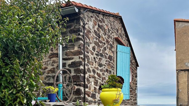 Un edificio de piedra con una puerta azul, una maceta amarilla en la pared y vistas al mar.