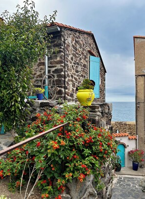 Ein Steinhaus mit einer blauen Tür, einem gelben Topf an der Wand und Blick auf das Meer.