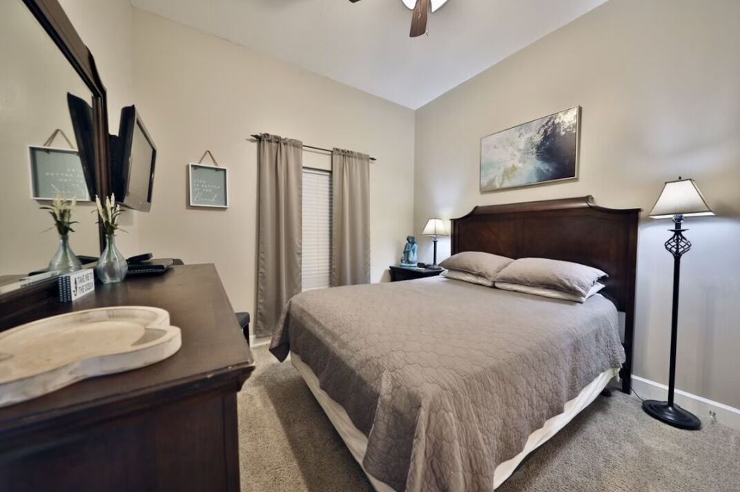 A bedroom with a bed, a desk with a mirror, a nightstand, a lamp, and a painting on the wall.