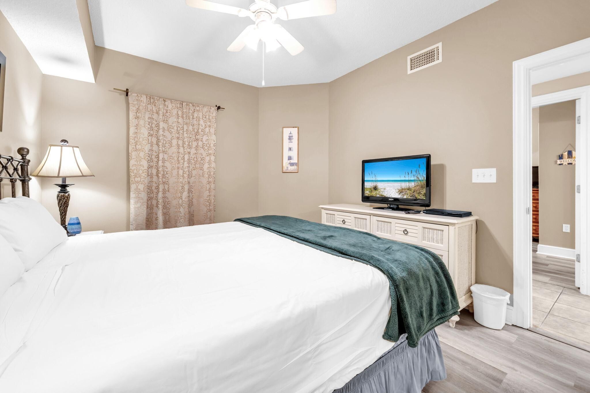 A bedroom with a bed, a television, a ceiling fan, a nightstand, and a door leading to another room.