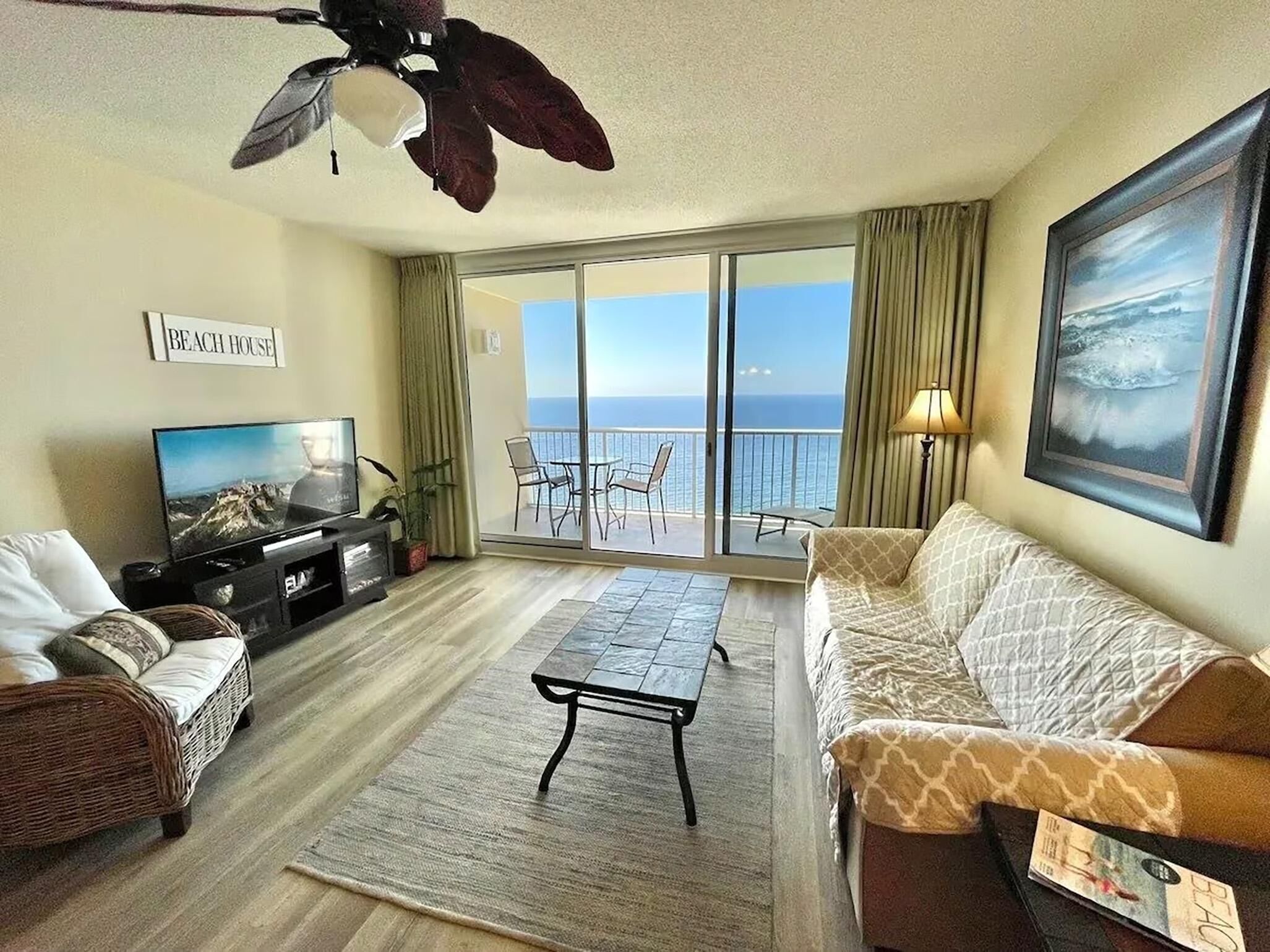 A living room with a television, sofa, and a balcony with a view of the ocean.