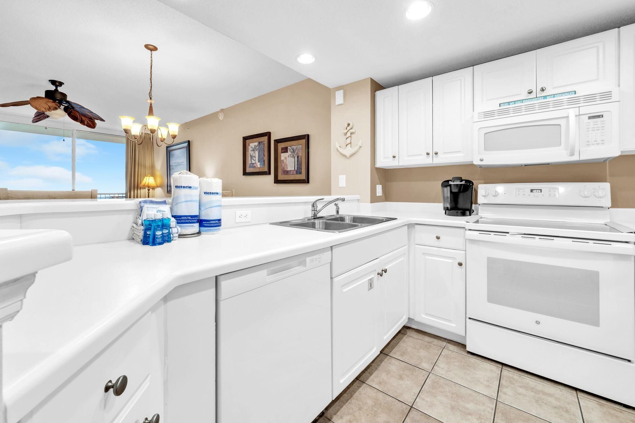 A modern kitchen with white cabinetry, a built-in microwave, a coffee maker, and a dishwasher. There are framed pictures on the wall and a ceiling fan.