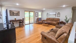 A spacious living room with a wooden floor, a brown sofa, a coffee table, and a TV on a stand. There is a dining area with a table and chairs, and a sliding glass door leading to a balcony. - 4BR/2BA with Huge Deck, Bonus Room, Swimming/Fishing & Stunning Seasonal Views! (Mountain Home)