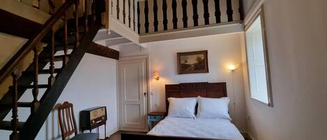A bedroom with a wooden bed, a chair, a nightstand, a radio, a painting, and a staircase.
