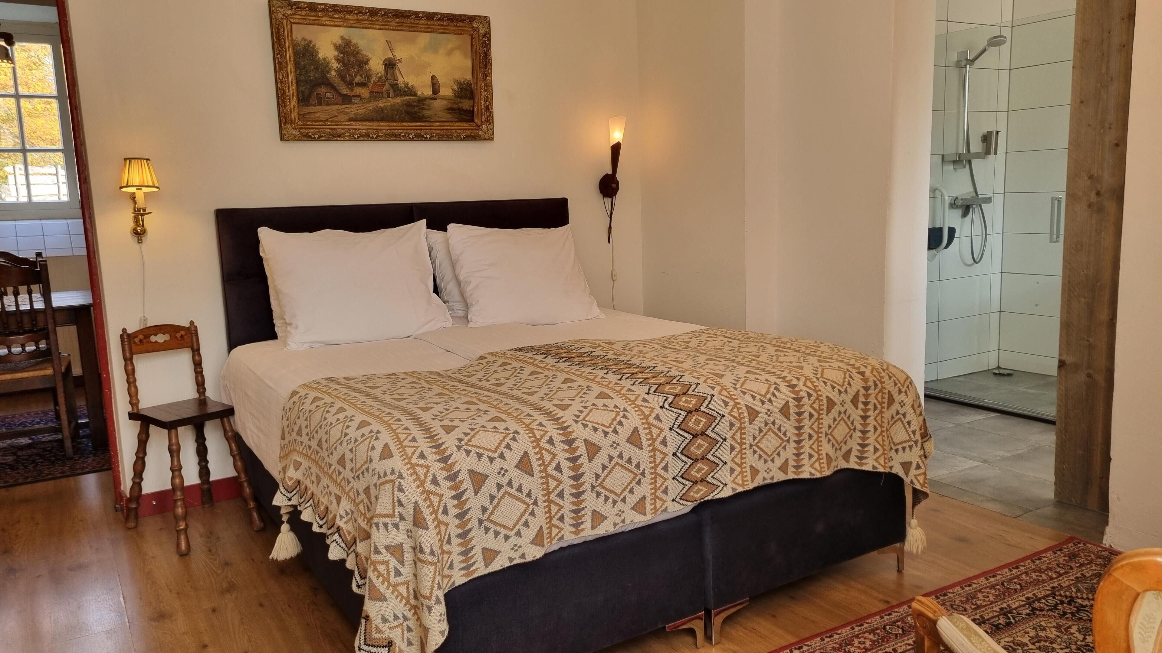 A bedroom with a bed, a nightstand, a chair, a painting, and a shower area.