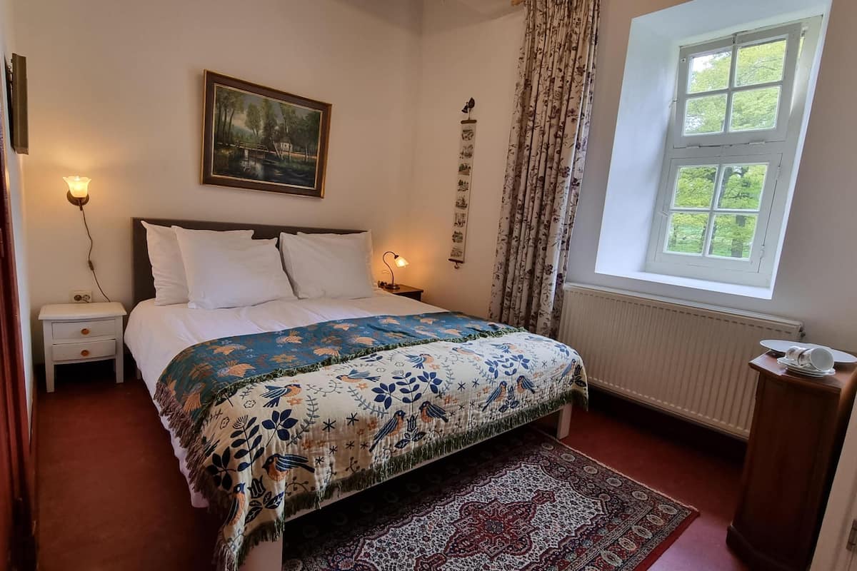 Une chambre à coucher avec un lit, deux tables de chevet, une commode, une fenêtre avec des rideaux, un radiateur et un tableau au mur.