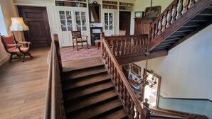 A grand staircase with wooden balusters and a chandelier, leading to a landing with a fireplace and bookshelves.