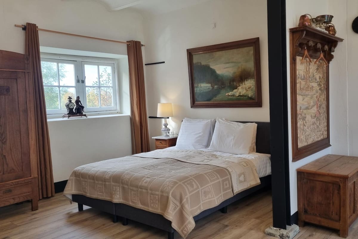 Une chambre à coucher avec un lit, une tête de lit en bois, une table de chevet avec une lampe, une fenêtre avec des rideaux et un tableau au mur.