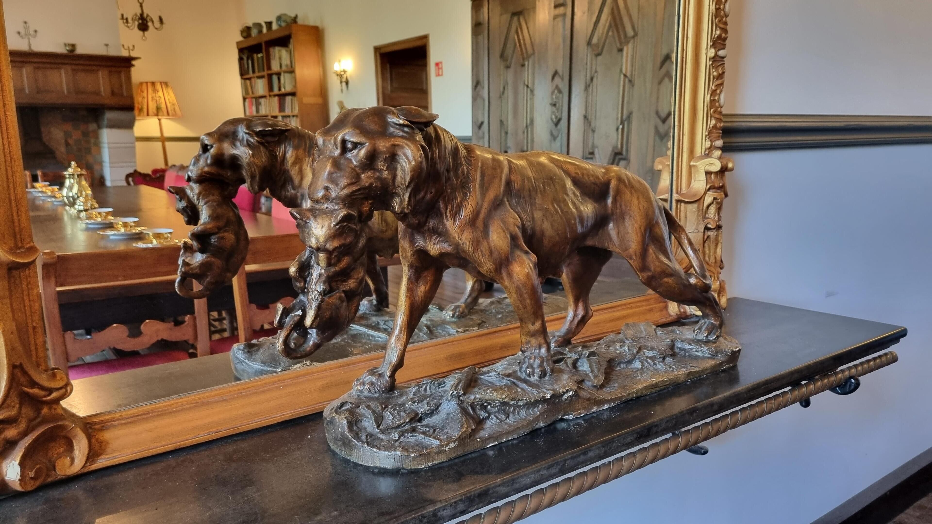 En træskulptur af en hund står på en hylde med et spejl, der reflekterer et rum med pejs, bogreoler og en dør.