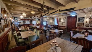 A cozy restaurant with wooden tables and chairs, hanging lights, and a bar area in the background.