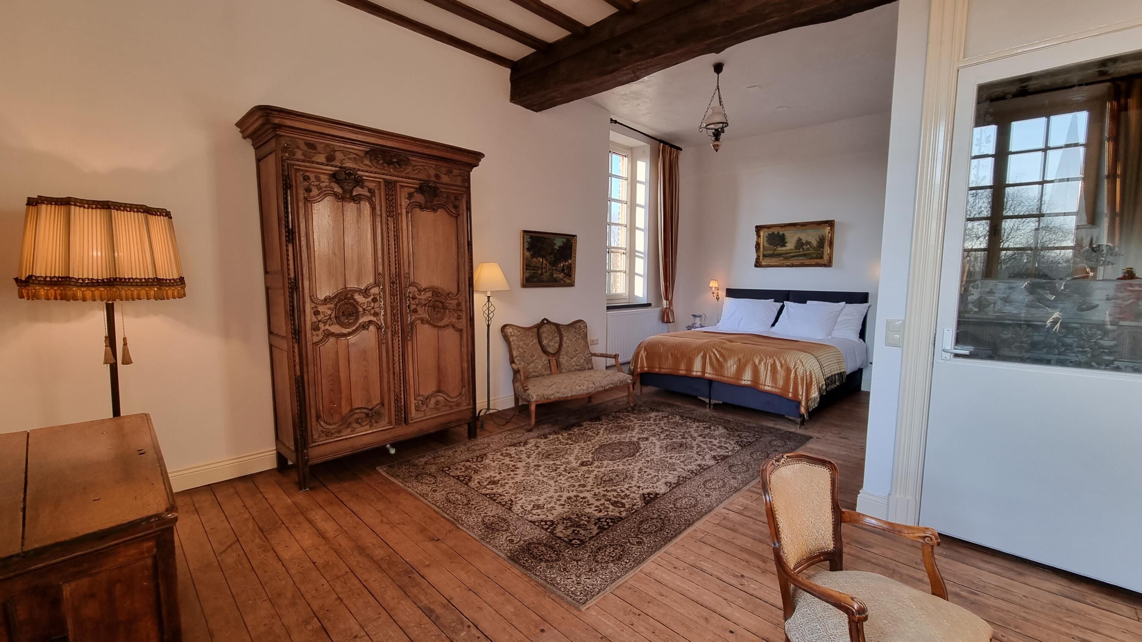 Un dormitorio con un armario de madera, una cama con colcha estampada, una silla, una lámpara de pie y una ventana con cortinas.